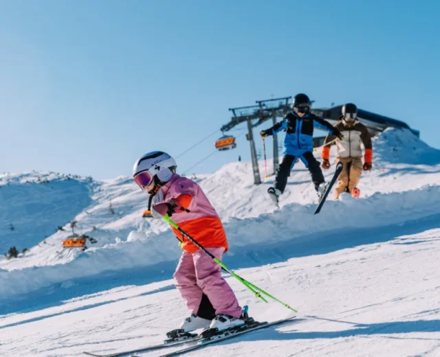 Dziecko w różowych nartach zjeżdża z góry we Flachau ferie z rodziną, dorośli podążają pod czystym, błękitnym niebem.