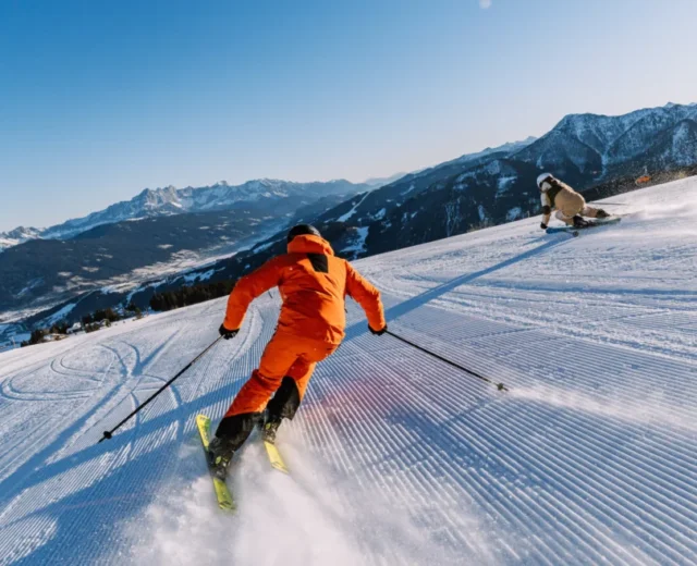 Dwie osoby zjeżdżają na nartach pod czystym, błękitnym niebem, ciesząc się nartami we Flachau i oszałamiającymi ośnieżonymi górami.