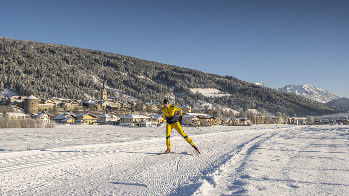 Narciarz na biegówkach w zółtym kombinezonie sunie po przetartej trasie stylem dowolnym. W tle zbocza gór i miasteczko Radstadt z wieżą kościoła i murami miejskimi.