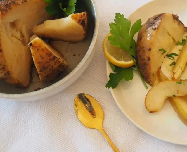 Seler pieczony w plastrach podawany z natką pietruszki, cytryną i złotą zastawą na białym obrusie.
