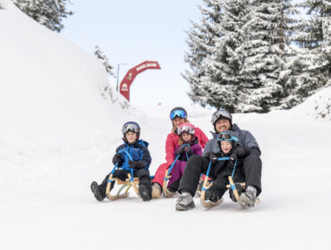 Czteroosobowa rodzina jeżdżąca na sankach w Radstadt zima, wszyscy uśmiechnięci i ubrani w sprzęt zimowy i kaski.