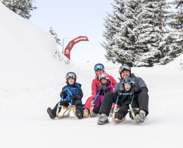Rodeln @ Lorenz Masser (2) Czteroosobowa rodzina jeżdżąca na sankach w Radstadt zima, wszyscy uśmiechnięci i ubrani w sprzęt zimowy i kaski.