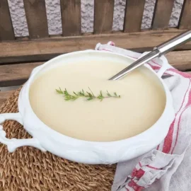 Biała waza z aksamitną zupą selerową, ozdobioną gałązką rozmarynu na słomiance, obok biała ściereczka w czerwone wzory.