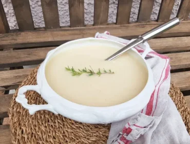 shared_recipe_1729516122.jpg Biała waza z aksamitną zupą selerową, ozdobioną gałązką rozmarynu na słomiance, obok biała ściereczka w czerwone wzory.