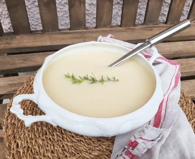 Biała waza z aksamitną zupą selerową, ozdobioną gałązką rozmarynu na słomiance, obok biała ściereczka w czerwone wzory.