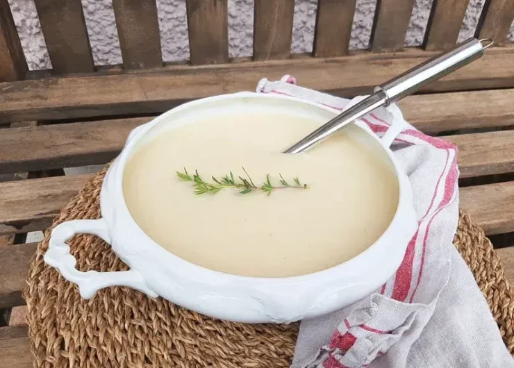 Biała waza z aksamitną zupą selerową, ozdobioną gałązką rozmarynu na słomiance, obok biała ściereczka w czerwone wzory.