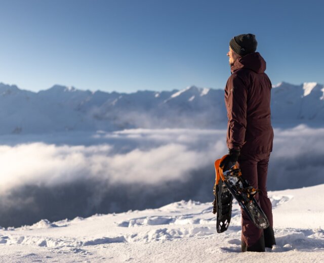 Osoba w zimowym stroju stoi na ośnieżonej górze, trzymając rakiety śnieżne, patrząc na odległe ośnieżone szczyty i chmury.