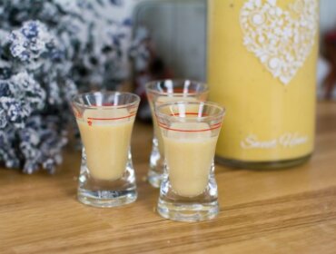 Trzy kieliszki do kremowego likieru jajecznego na drewnie, obok świąteczna butelka i kwiaty.