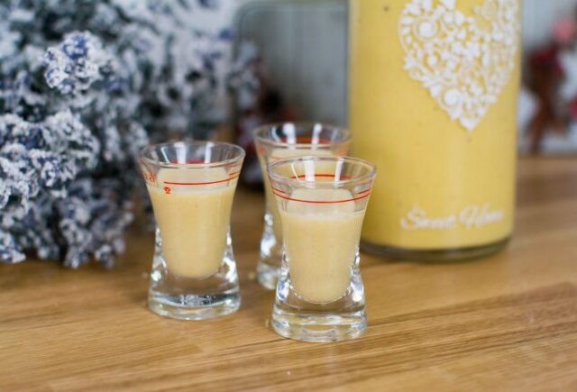 Trzy kieliszki do kremowego likieru jajecznego na drewnie, obok świąteczna butelka i kwiaty.