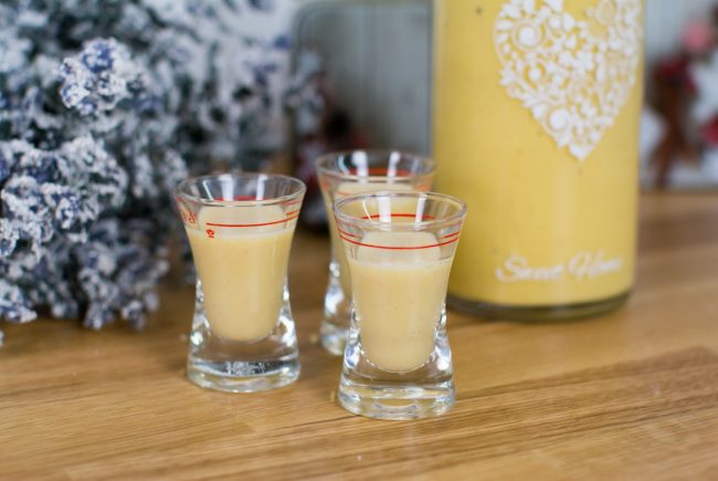 Trzy kieliszki do kremowego likieru jajecznego na drewnie, obok świąteczna butelka i kwiaty.