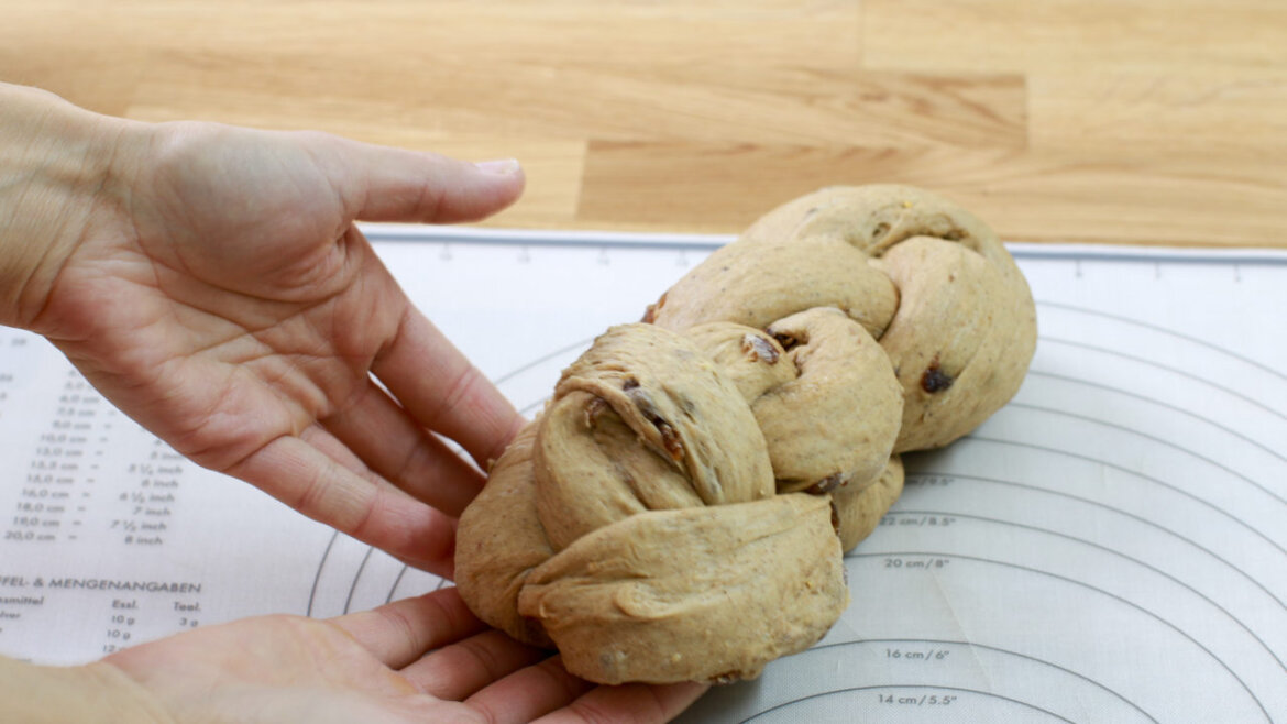 Krok 12 © Anke Eder Dwie ręce trzymające spleciony bochenek ciasta na macie do pieczenia z oznaczeniami pomiarowymi. (Powiększony widok)