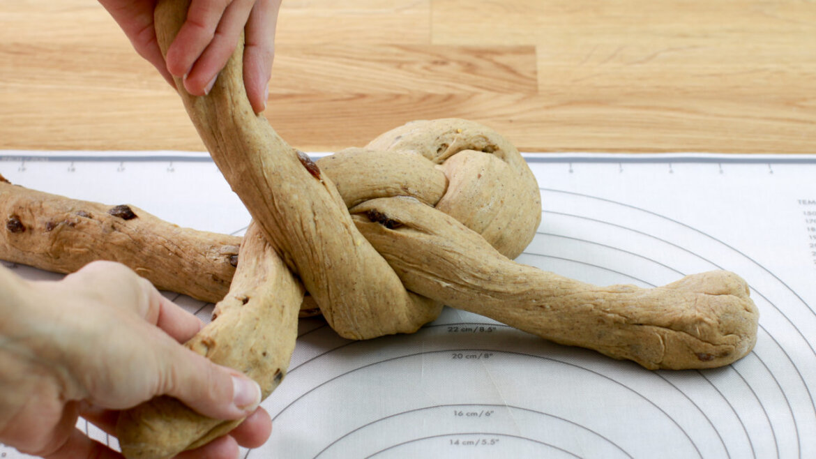 Krok 9 © Anke Eder Ręce zaplatające ciasto na macie do ciasta z prowadnicami pomiarowymi, na drewnianym blacie. (Powiększony widok)