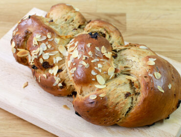 Fruehstuecksstriezel-fuer-Weihnachten-2048×1365 Pleciony bochenek słodkiego chleba z rodzynkami i pokrojonymi migdałami na drewnianej desce do krojenia.