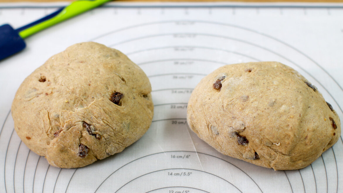 Dwie kulki ciasta chałkowego z rodzynkami na macie do ciasta, z niebieską i zieloną szpatułką w tle.