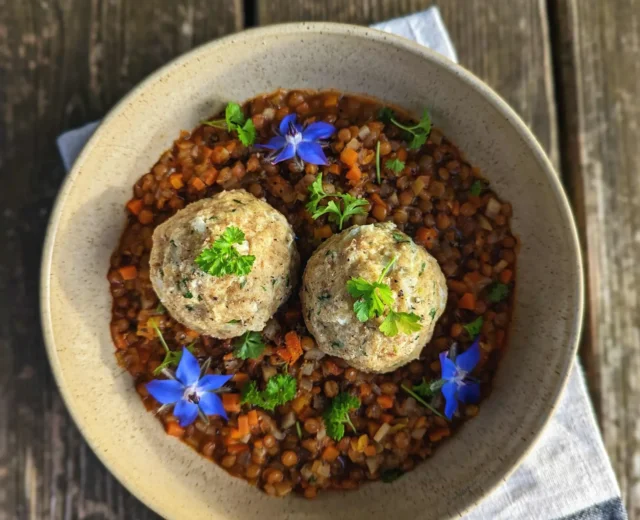 Berglinsen mit Wurzelgemüse und Semmelknödel_Isstgut_Höfelmaier.jpg Miska gulaszu z soczewicy z dwoma pierożkami, świeżymi ziołami i niebieskimi jadalnymi kwiatami na serwetce.