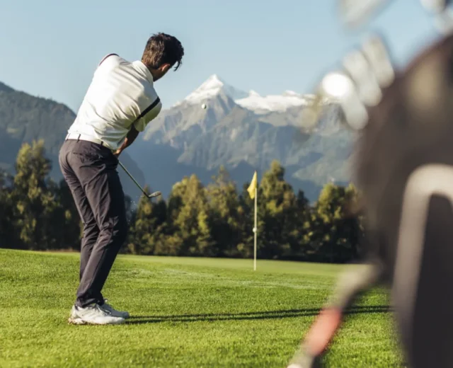golfen-mit-blick-aufs-kitzsteinhorn_c-zell-am-see-kaprun-tourismus-b82c0823_medium-1170×658.jpg Golfista zamachuje się w kierunku greenu z górami i drzewami w tle w słoneczny dzień.