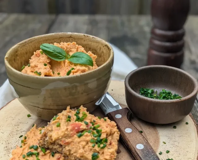 Linsenaufstrich mit Tomaten I_web©Salzburger Agrar Marketing_Martina Höfelmaier.jpg Miseczka z pastą pomarańczową, nóż i kromki chleba z pastą i szczypiorkiem na drewnianej desce.