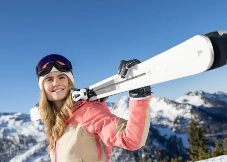 Uśmiechnięta kobieta w sprzęcie narciarskim trzymająca narty na ramieniu z ośnieżonymi górami w tle.