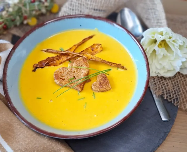 shared_recipe_1716528615.jpg Miska kremowej żółtej zupy z grzankami, smażonymi paskami i szczypiorkiem, obok białego kwiatka i łyżki.