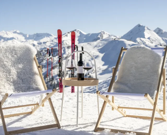 ski-und-weingenusswoche-skigebiet-8 Dwa krzesła i mały stolik z winem na ośnieżonych górach, narty w tle pod czystym błękitnym niebem.