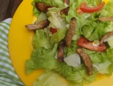 Sałatka z sałatą, pomidorem, grillowanymi paskami mięsa i jadalnymi kwiatami na żółtym talerzu na drewnianym stole.